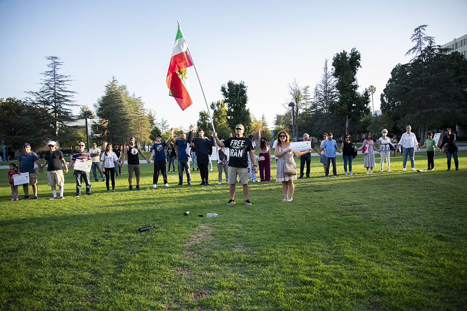 People stand on the University Library lawn, many holding hands with an Iranian flag.