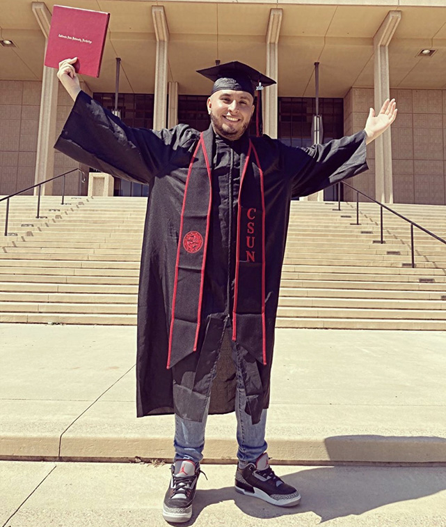 A male student in graduation cap and gown holds up his arms in triumph in front of the CSUN Library.