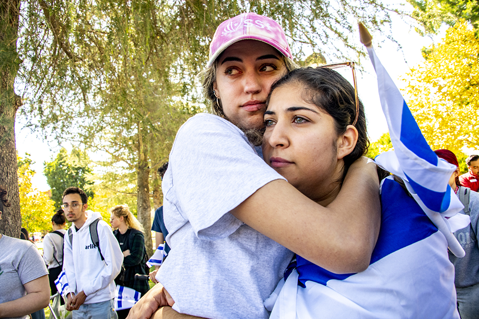 Two women stand with their arms around each other. One woman wears the blue and white Israeli flag around her shoulders.