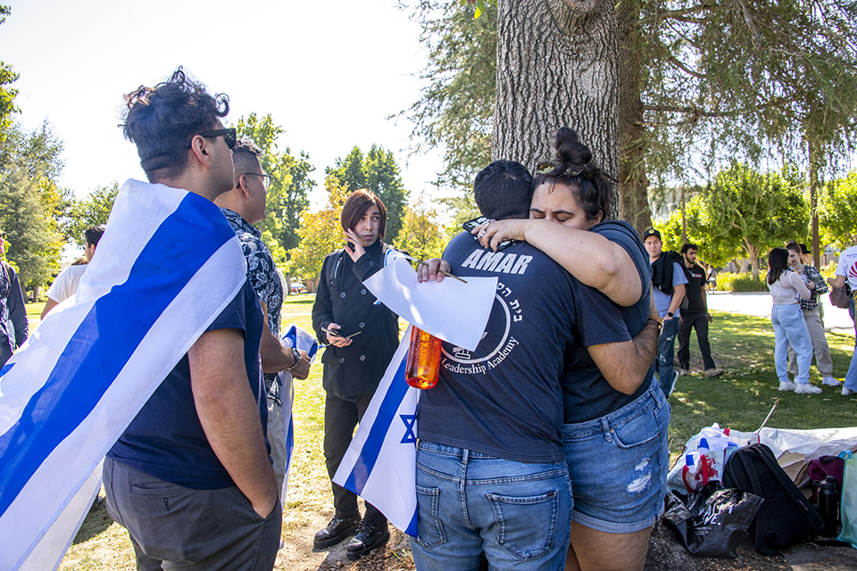 Two students stand by a tree and hug each other, while another student wearing an Israeli flag, looks on.