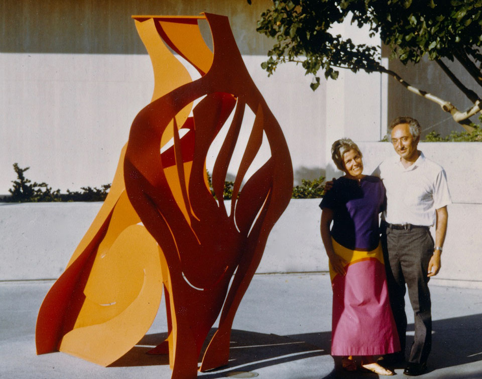 Artists Irene Monat Stern and Jan Peter Stern stand next to one of his vibrant orange, painted-steel sculptures.