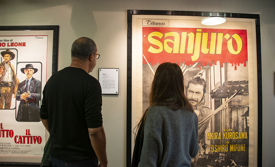A man and a woman look at framed, vintage film posters by legendary filmmakers Akira Kurosawa and Sergio Leone.
