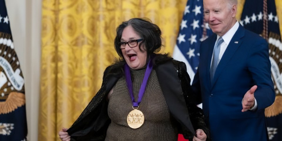 Judy Baca shows off her National Medal of Arts. President Biden stands to the side of her.