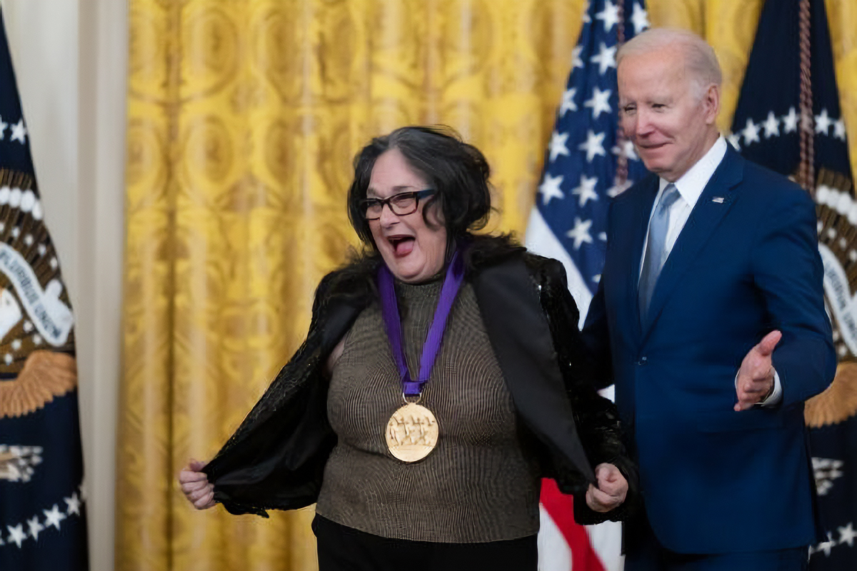 Judy Baca shows off her National Medal of Arts. President Biden stands to the side of her.