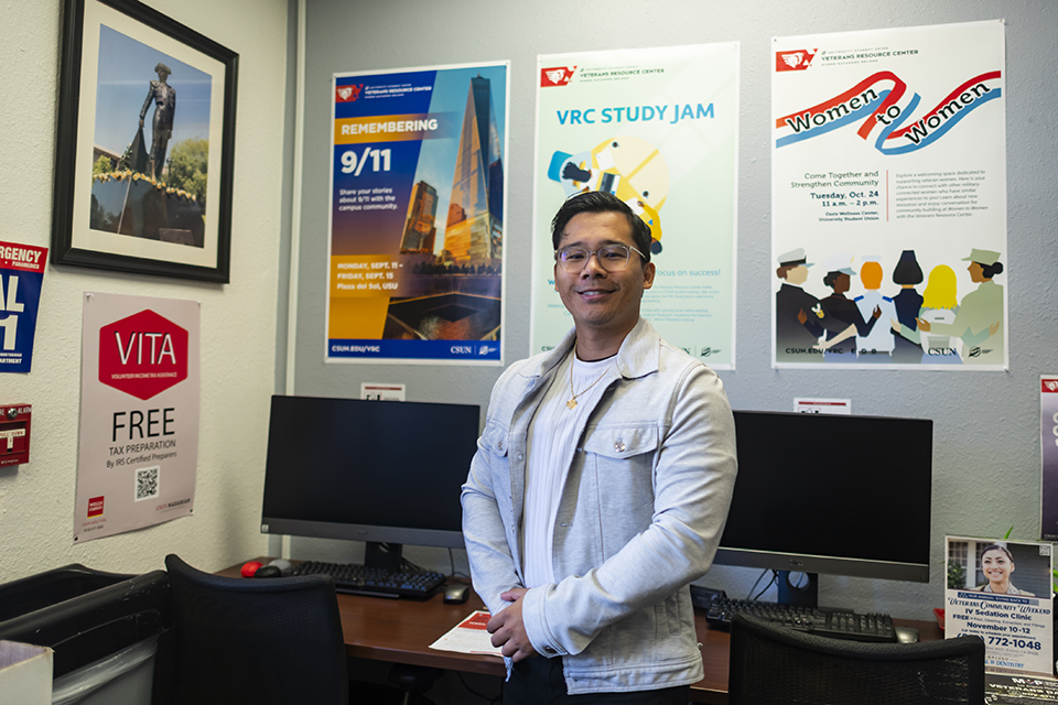 Justin Aragon stands in front of posters showing services provided by the Veterans Resource Center