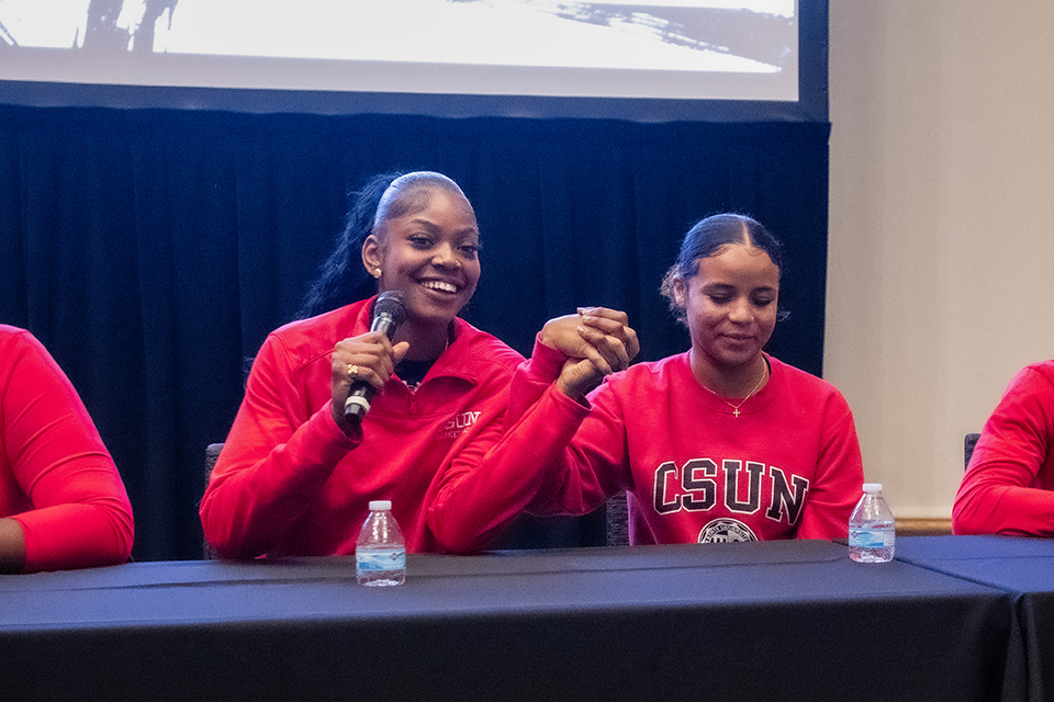 Two women wearing red team shirts sit at podium and hold their hands together.