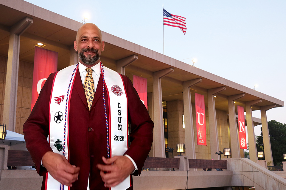 A male student poses in a suit and graduation sash in front of a virtual CSUN Library background.