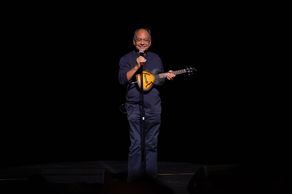 Comedian Cheech Marin, holding a ukulele, is spotlit against an all-black background.