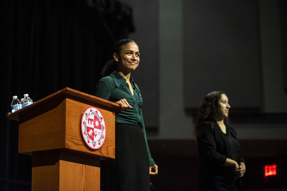 Lauren Ridloff stands smiling on stage by podium with CSUN seal. An American Sign Language translator, dressed in black stands in the background.