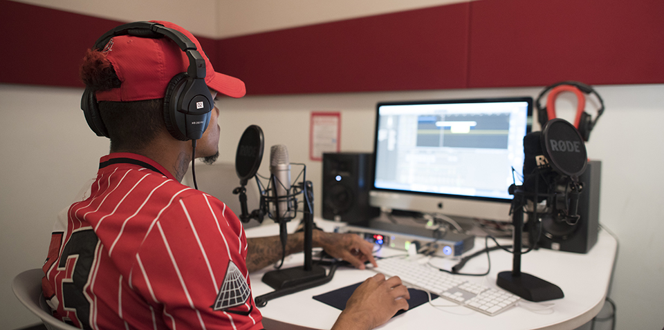 Student wearing headphones uses equipment in the Audio Recording Studio. He uses a keyboard to edit his audio, and has two microphones on the desk.