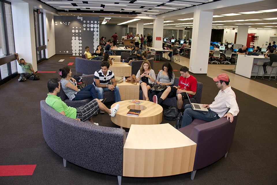 Students sit at a round lounge sofa while talking, using their phones and studying.