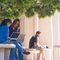 Two students sit on a bench with their laptops open in front of the University Library