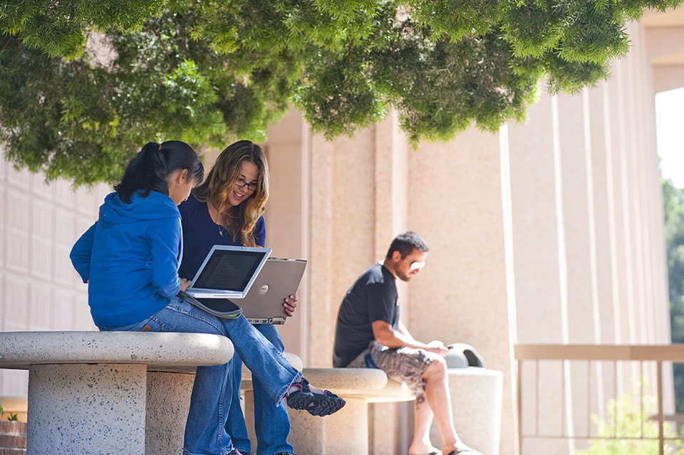 Two students sit on a bench with their laptops open in front of the University Library