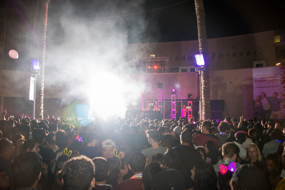 Crowd of students outdoors in front of stage with DJs and bright lights.