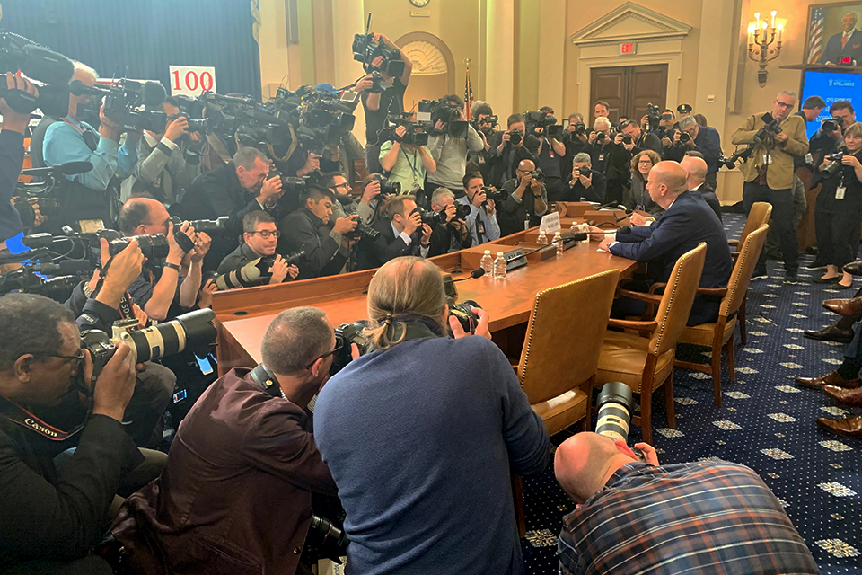 Politicians at a desk are surrounded by a crowd of reporters and photographers.