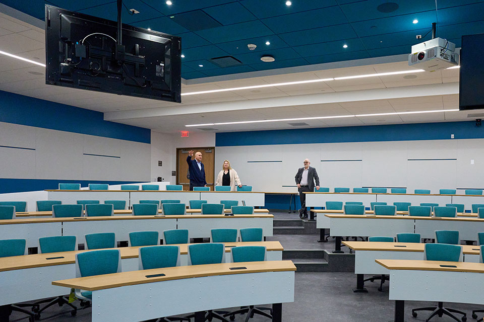 Horizontal photo of large lecture room with rows of desks and blue chairs. In the back of the room, President Erika Beck is standing with Ken Rosenthal and Colin Donahue, looking over the room.
