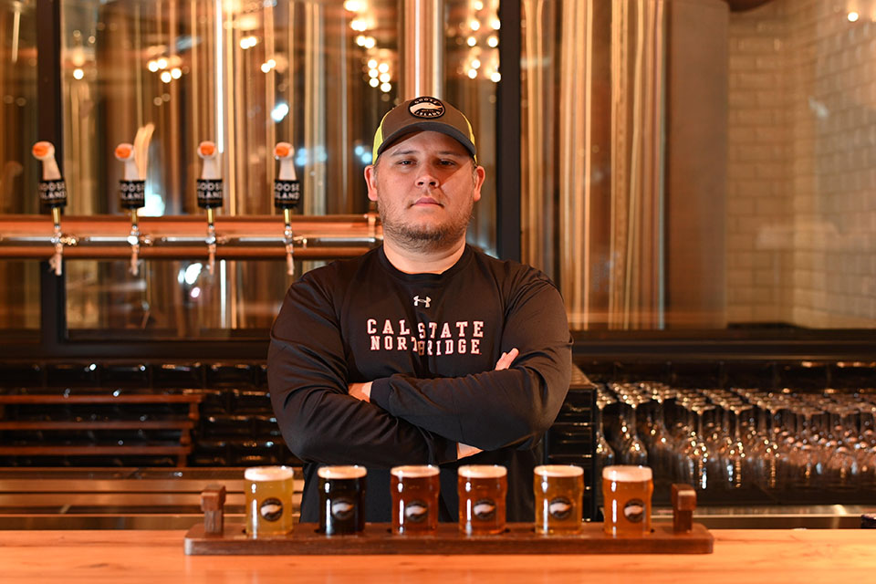 Alumnus Matt Jimenez stands behind a bar in a Goose Island tasting room, in front of a poured flight of Goose Island beers.