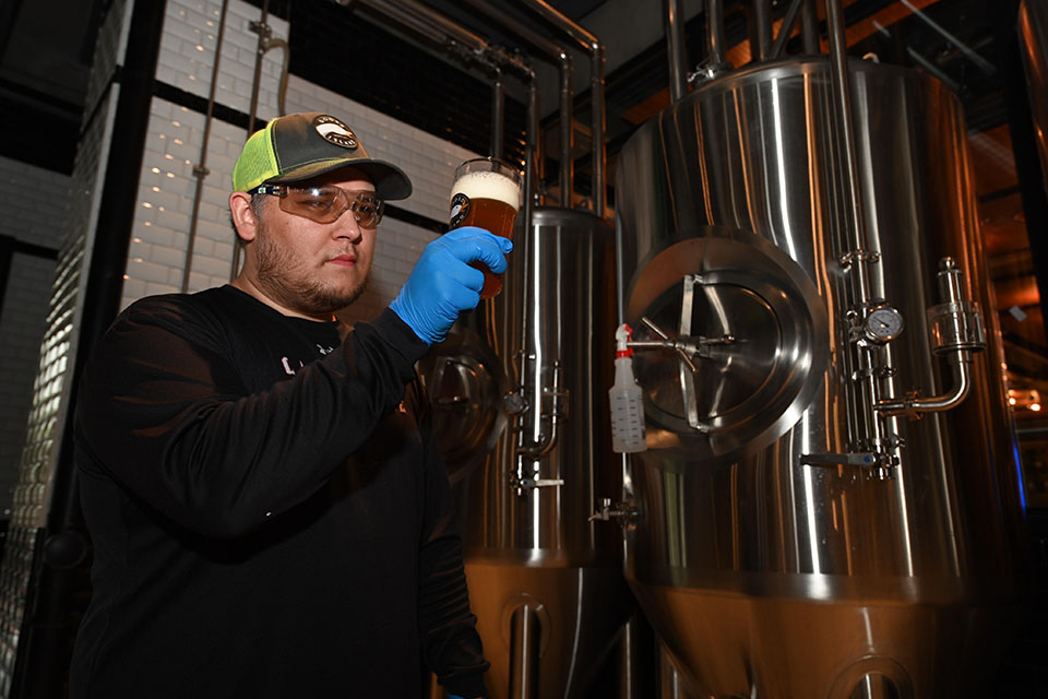 Alumnus Matt Jimenez, wearing a Goose Island baseball cap, protective goggles and gloves, holds up a pint of beer, examining the color, clarity and foam.