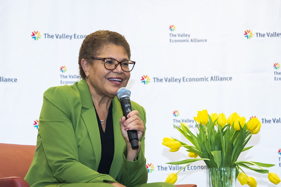 Los Angeles Mayor Karen Bass sits on stage, holding a microphone and speaking to an audience.