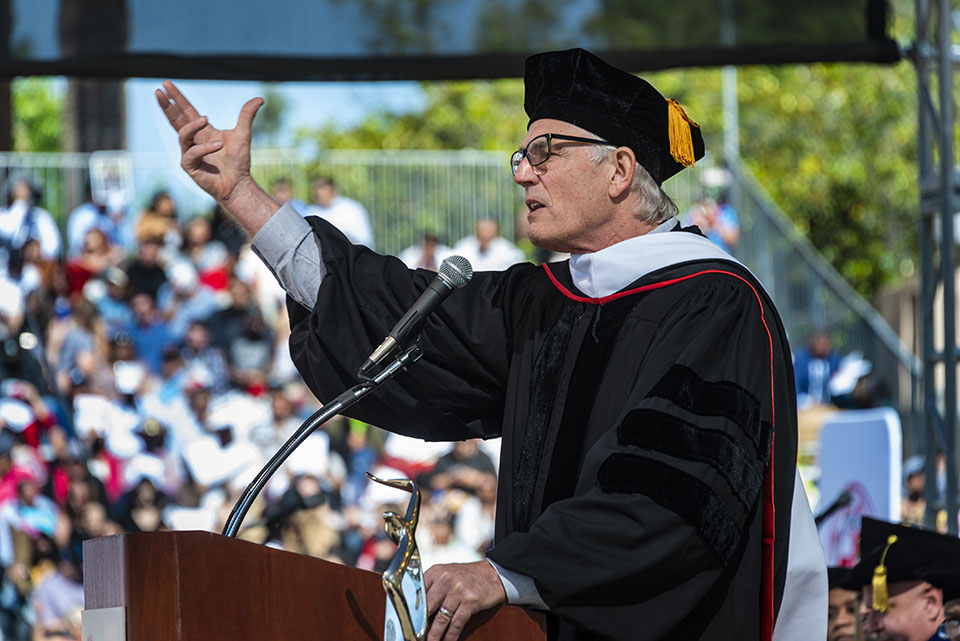 Honorary Doctorate recipient and CSUN alumnus stands a podium, raising his hand toward the sky and speaking to graduates at Commencement 2022.