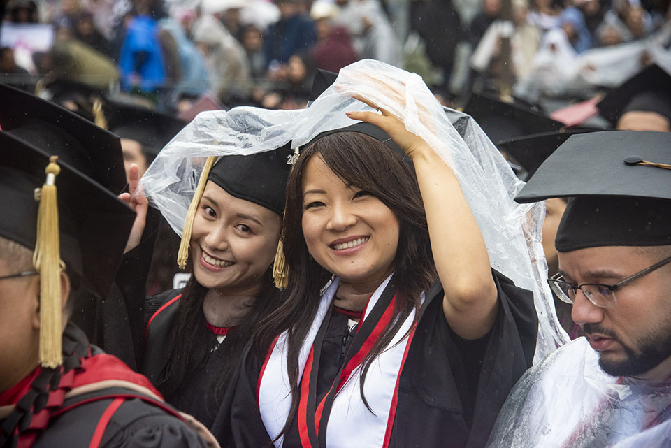 Two college graduates share a plastic raincoat to cover themselves from the rain during the 2019 commencement ceremony.