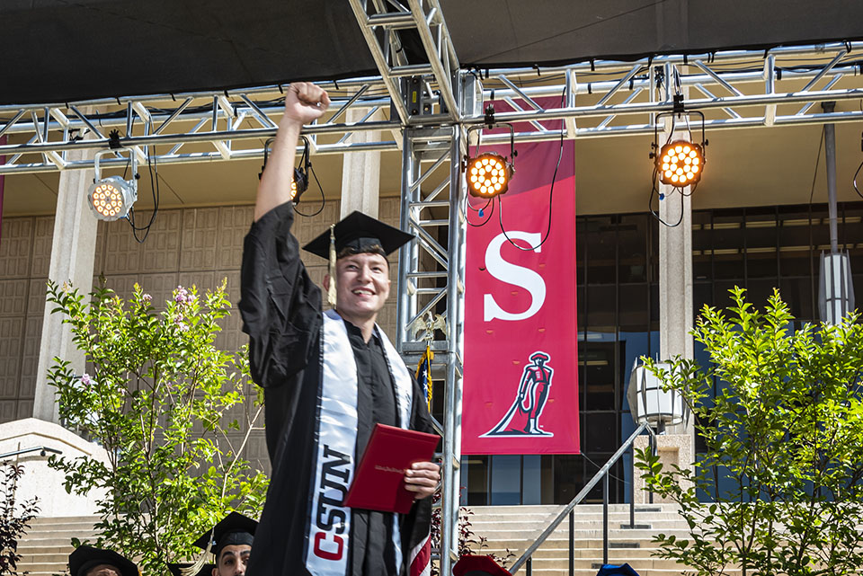 A student-athlete and new graduate raises his fist in the air and holds his red diploma cover, as he crosses the stage at Commencement 2022.