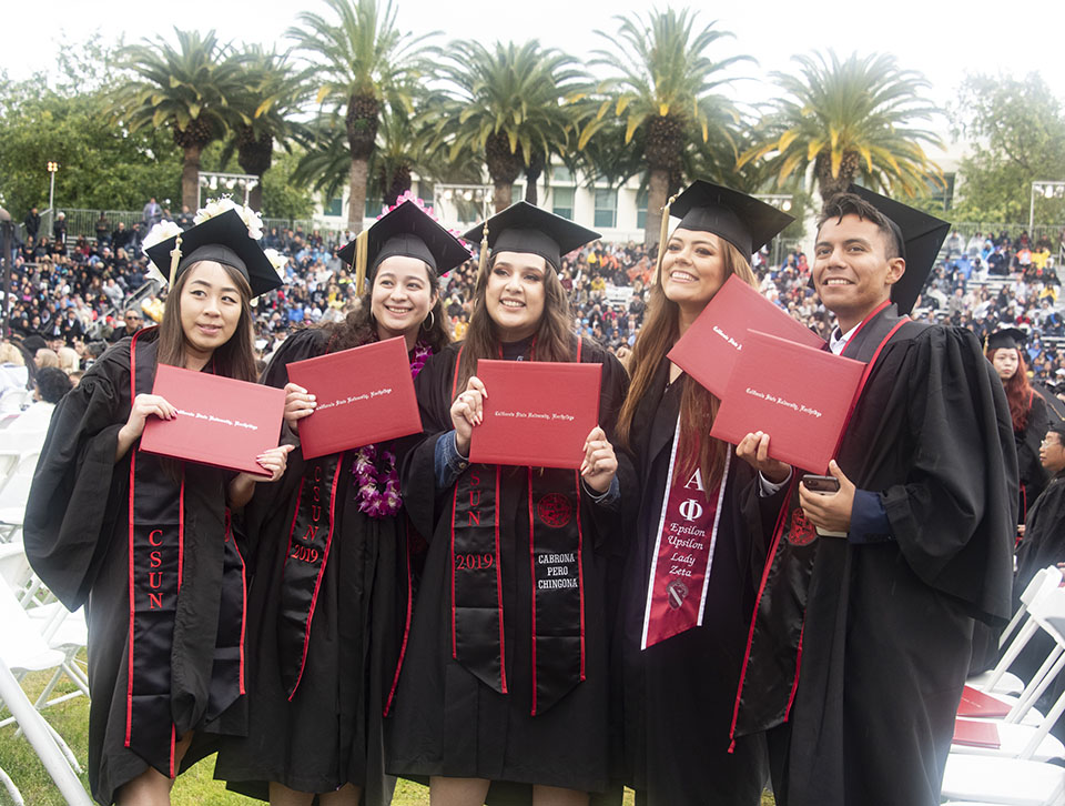 Group of five college graduates hold up their red diplomas and smile.
