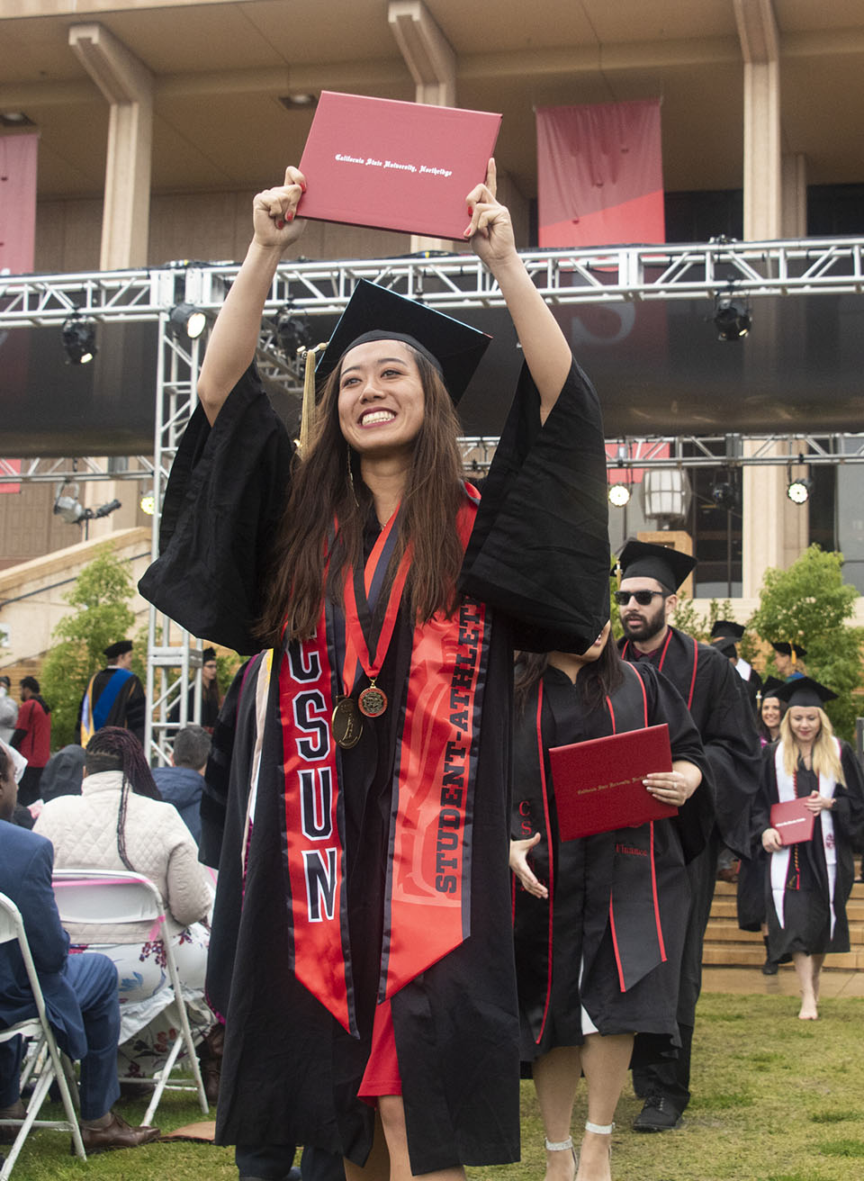Smiling college graduate is wearing a red stole that says 'CSUN student athlete' and is holding up a red diploma.