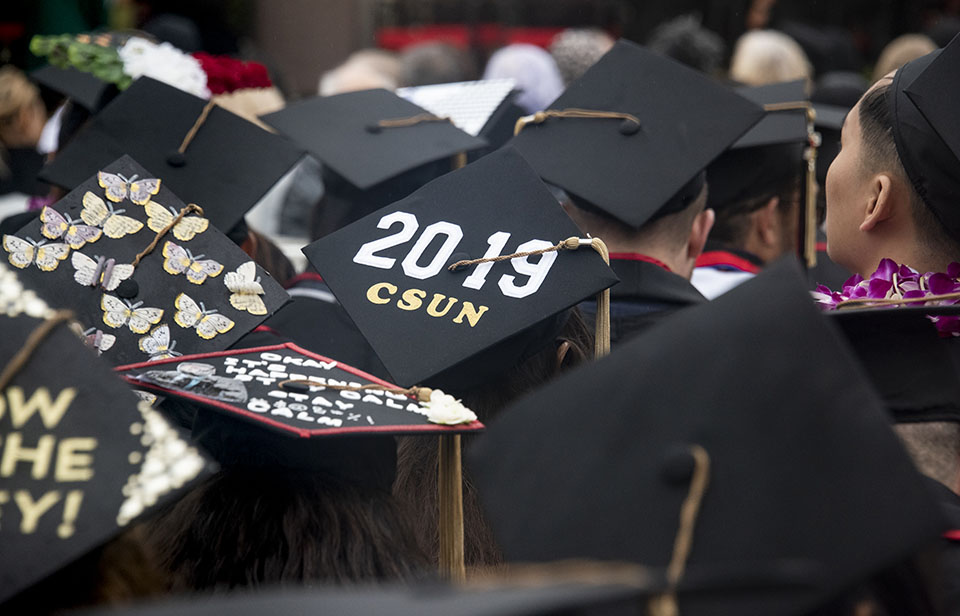 Black graduation cap that says 2019 CSUN on it is seen in the middle of a crowd of seated college graduates.