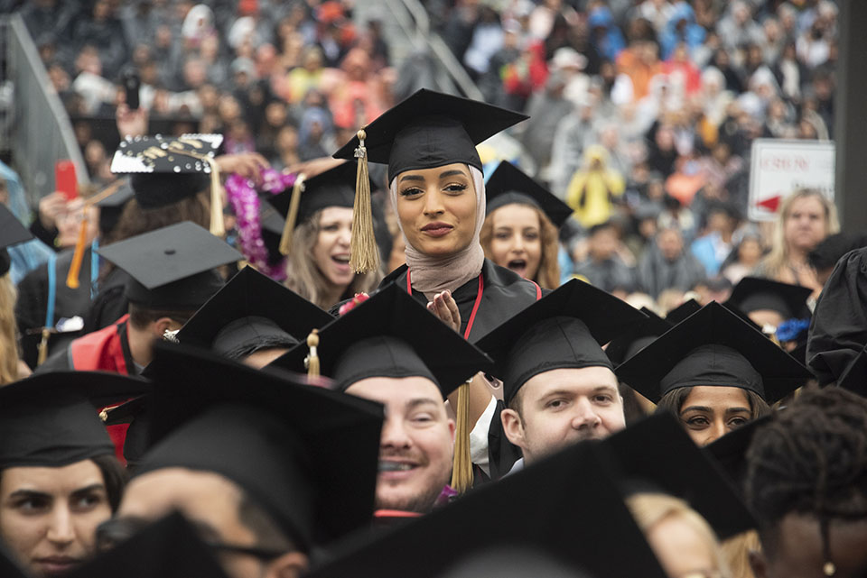 A female college student stands and claps in the middle of seated graduates.