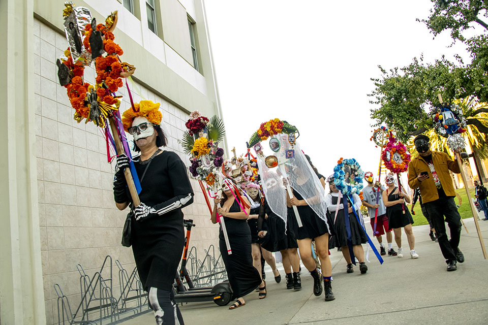 A procession of students and faculty members, wearing masks and makeup, carries colorful flower wreaths and banners for Noche de Ofrendas.