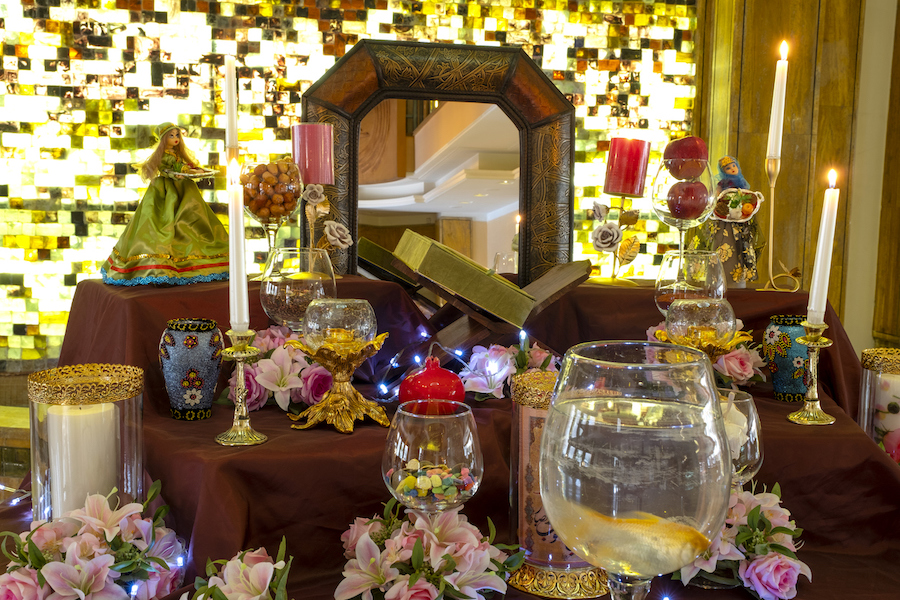 A traditional Persian New Year table set with a mirror in the center surrounded by white and red candles, colorful candies, fruit, pink flowers and goldfish.