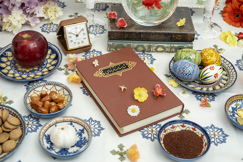A book with farsi print on the front sits at the center of the Haft Seen table. The book is surrounded by dishes filled with candy, spices and painted eggs.