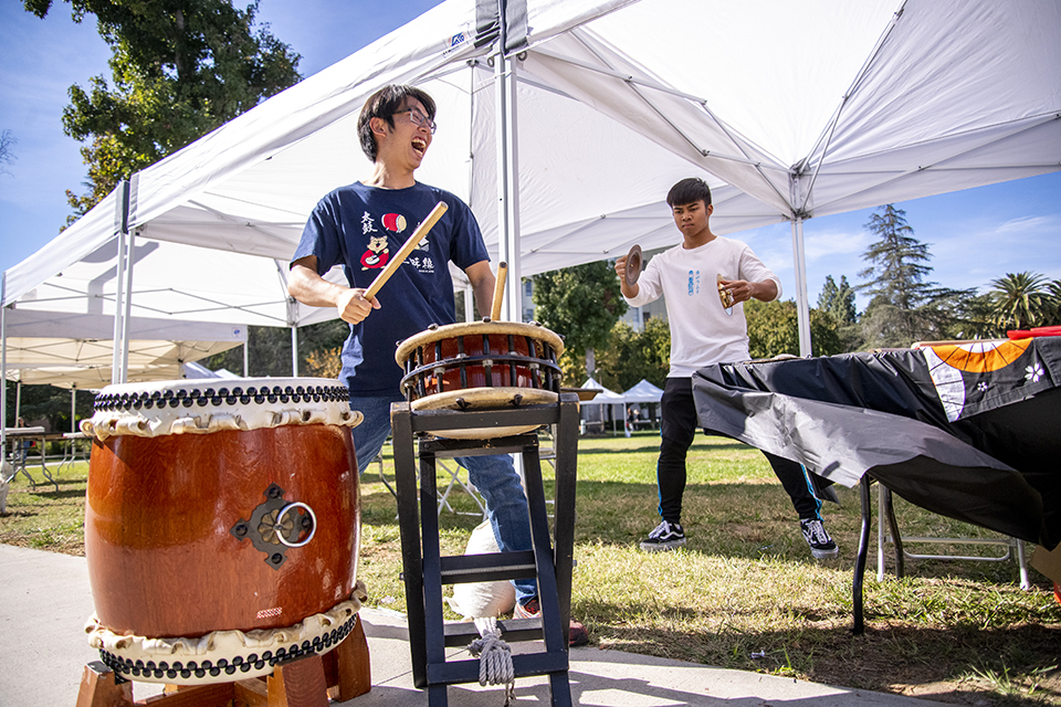 Young man smiles as he plays a large Japanese drum. Another, larger wooden drum stands beside him.