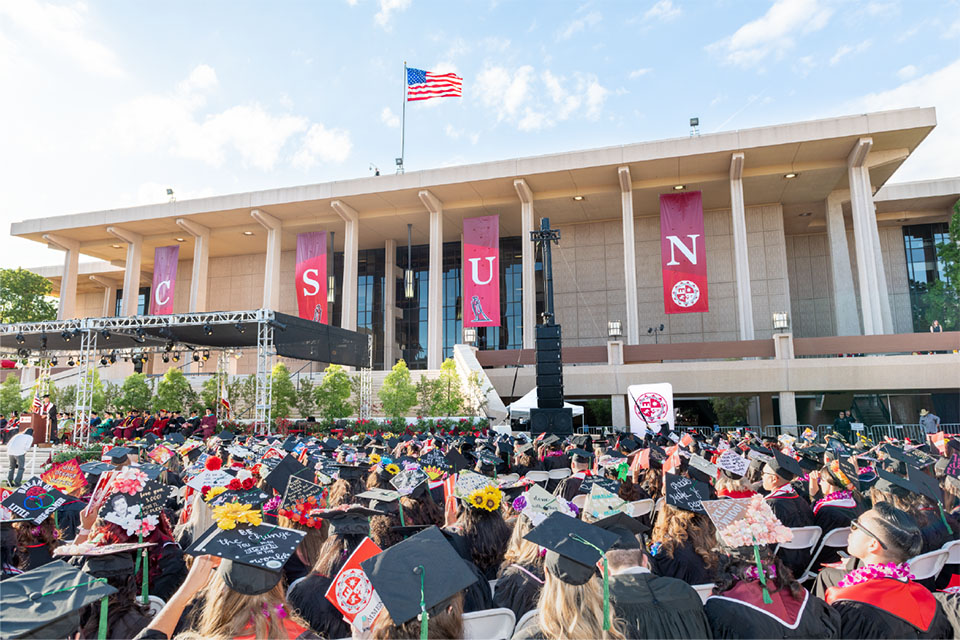 A crowd of CSUN graduates in caps and gowns sits in chairs in front of the Oviatt Library, with CSUN banners hanging and the American flag flying on the roof of the library.