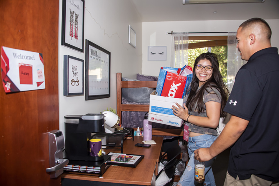 A student carries a box holding her belongings as she and a family member decorate her dorm room.