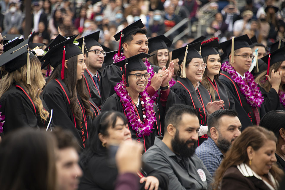 A crowd of CSUN graduates in caps, gowns and flower leis, stands and claps during Commencement ceremony.