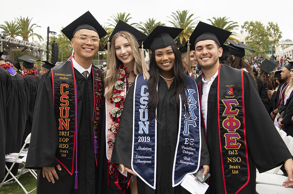 Four CSUN graduates in caps, gowns and sashes, smile and stand together at their seats in front of the University Library, at Commencement 2022.