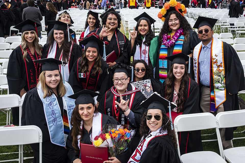 A crowd of CSUN graduates in caps, gowns and sashes, smile and show the ASL sign for 