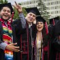 Three CSUN graduates in caps, gowns and sashes, smile and raise their arms together at their seats in front of the University Library, at Commencement 2022.