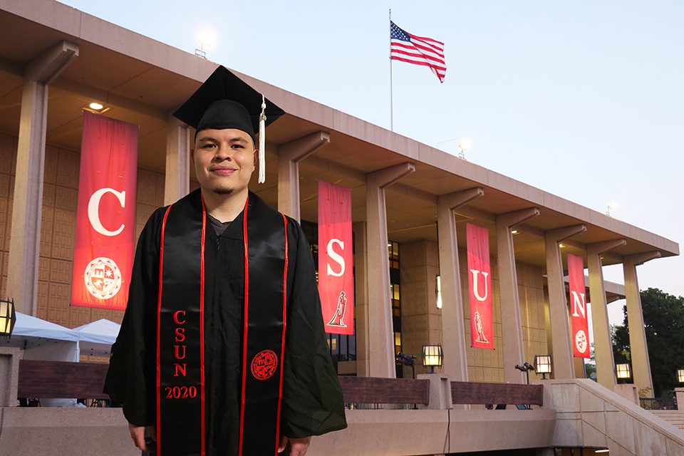 A male CSUN student poses in graduation cap and gown in front of a virtual CSUN Library background.
