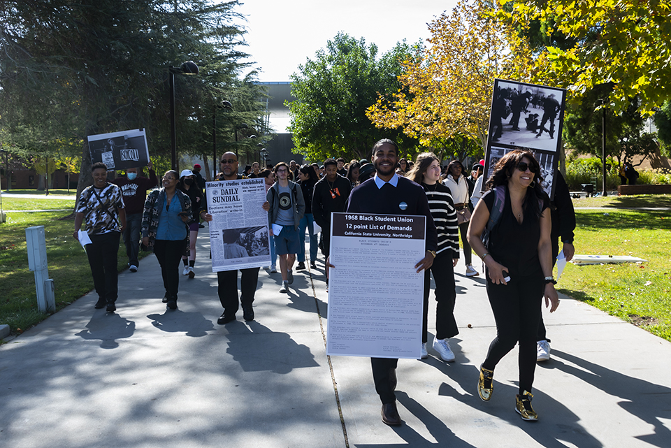 Students, staff, faculty and guests hold large signs made from the original 1960's protests while walking.