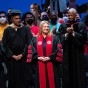President Erika D. Beck stands on stage at The Soraya, in a black and red CSUN academic gown, and flanked by President Emerita Jolene Koester, David Nazarian, Robert Taylor and CSU Acting Chancellor Steve Relyea. Behind her is a crowd of CSUN students in colorful T-shirts.