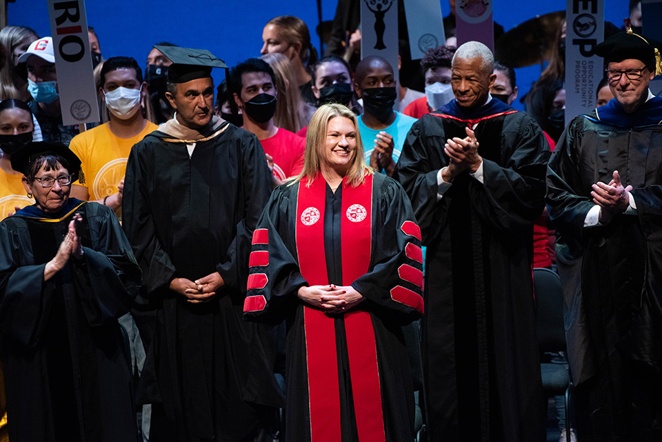President Erika D. Beck stands on stage at The Soraya, in a black and red CSUN academic gown, and flanked by President Emerita Jolene Koester, David Nazarian, Robert Taylor and CSU Acting Chancellor Steve Relyea. Behind her is a crowd of CSUN students in colorful T-shirts.