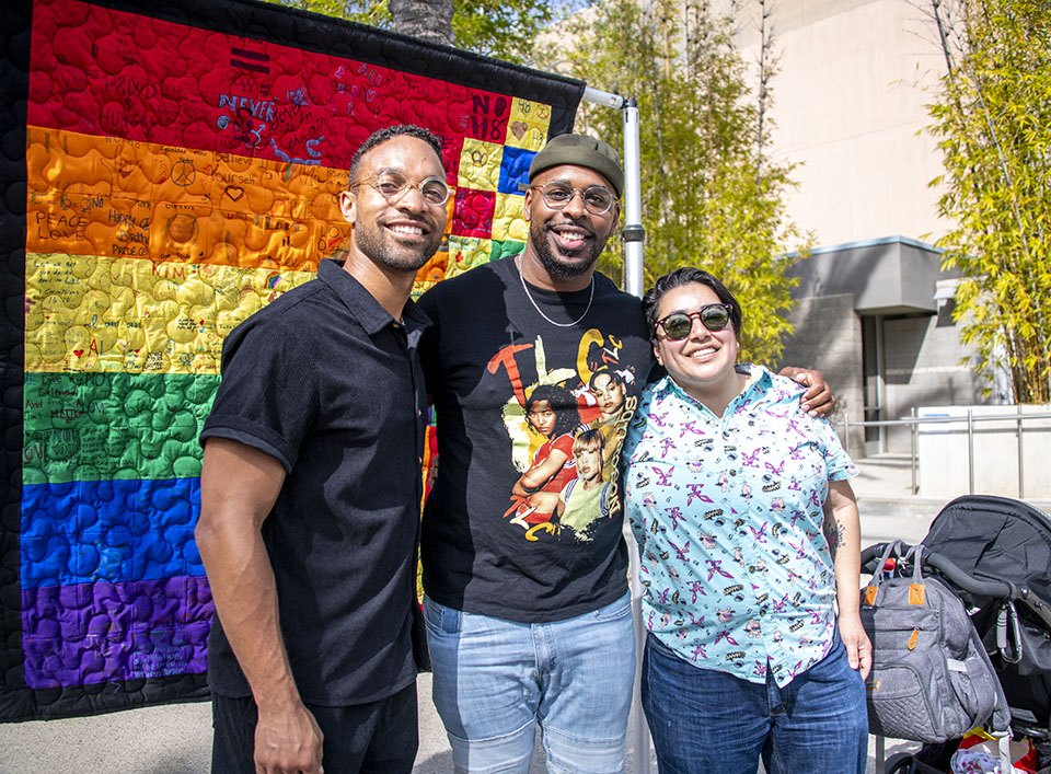 Three alumni stand in front of a quilted rainbow banner, signed by students who helped open the CSUN Pride Center in 2012.