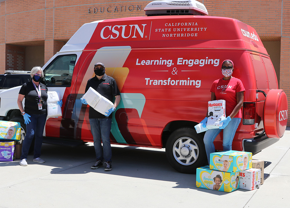 Three CSUN faculty and staff members stand in front of the red-and-white van for CSUN's Family Focus Resource Center, next to a pile of diaper boxes.