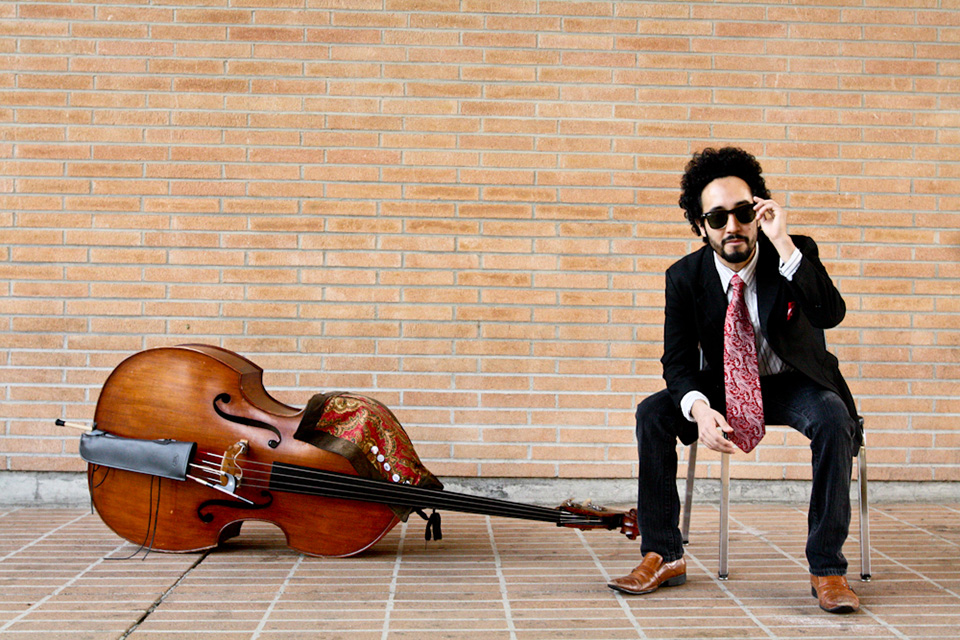 Musician Rodrigo Moreno sits in a chair, wearing sunglasses, next to his string bass.