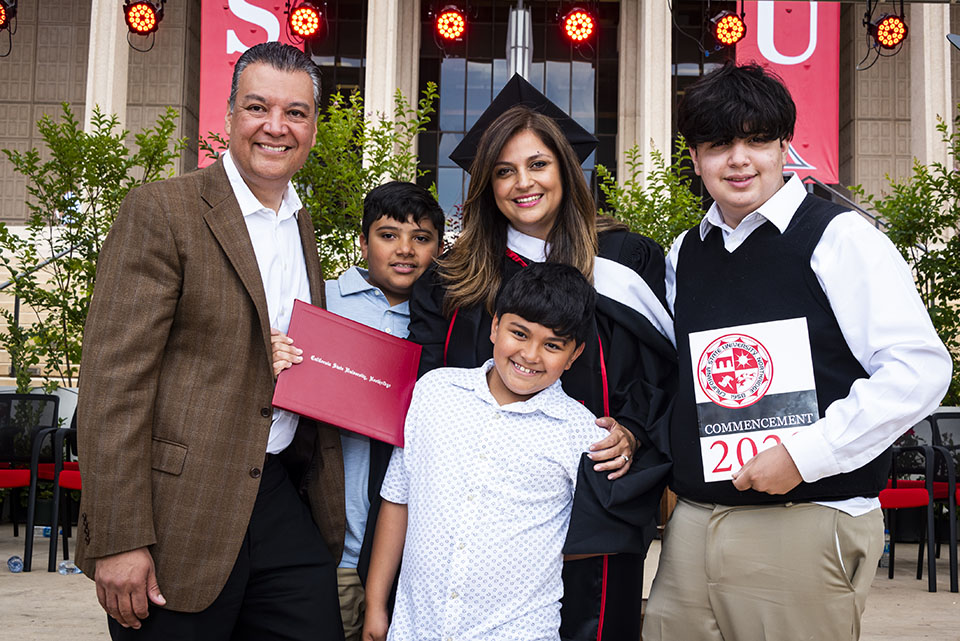 U.S. Senator Alex Padilla stands with his wife, Angela Gabriela Padilla (in cap and gown and holding her red diploma cover), with their three children, on stage after the Commencement ceremony for CSUN's College of Social and Behavioral Sciences.
