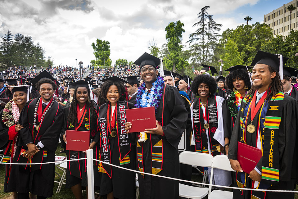 A crowd of graduates in caps, gowns and sashes smile and hold their red diploma covers, in front of the University Library at Commencement 2022.