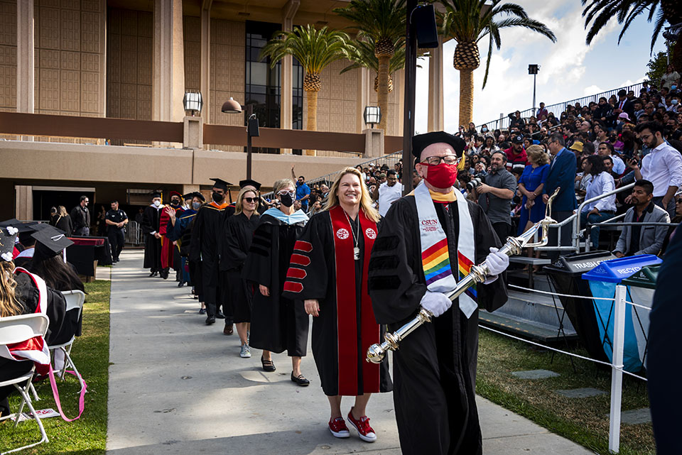 A faculty member carries the university mace and leads President Erika D. Beck and other CSUN leaders down a walkway in front of the University Library, at Commencement 2022.